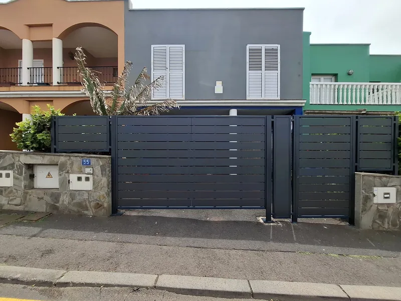 Acceso amplio gris de lamas horizontales y puerta peatonal a su derecha frente a una vivienda