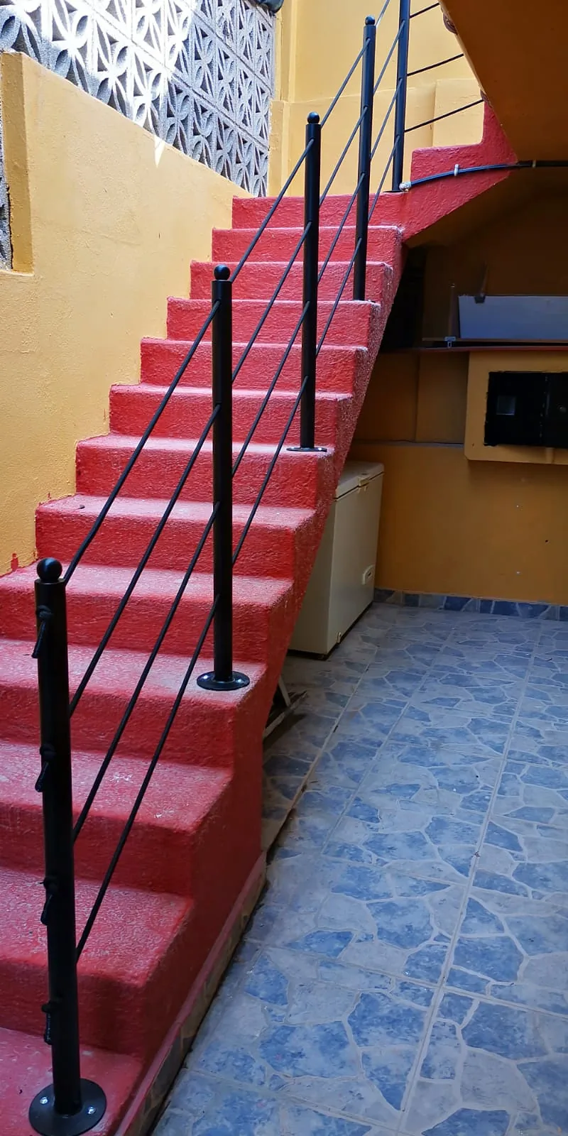 Barandilla metálica negra junto a una escalera roja de obra en un patio de paredes amarillas