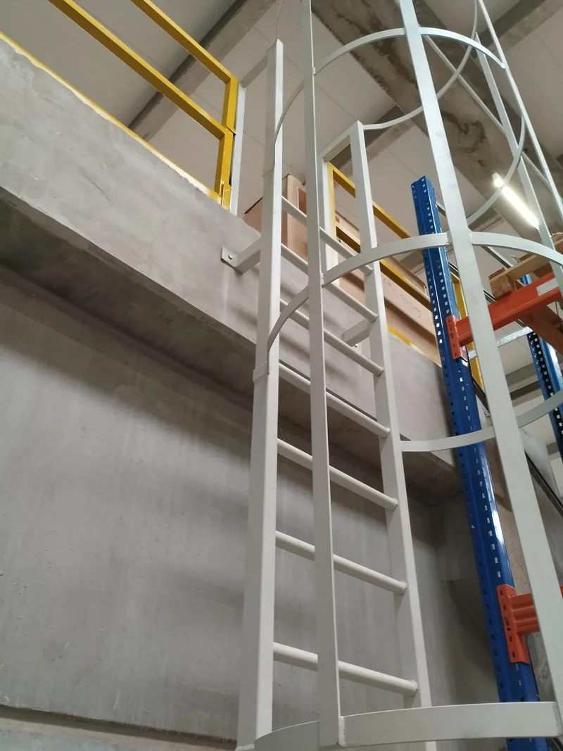 Escalera vertical blanca con aros y llegada al nivel de una barandilla amarilla en un almacén