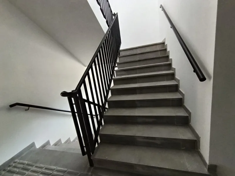 Tramo inclinado de barandilla negra y pasamanos tubular sobre una escalera interior de peldaños grises