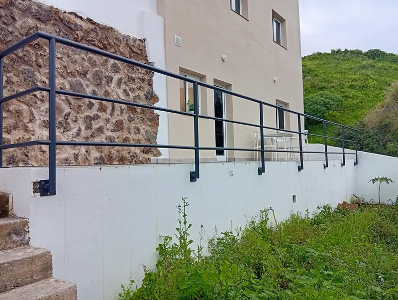 Barandilla tubular gris fijada al lateral de un muro blanco frente a una terraza de vivienda
