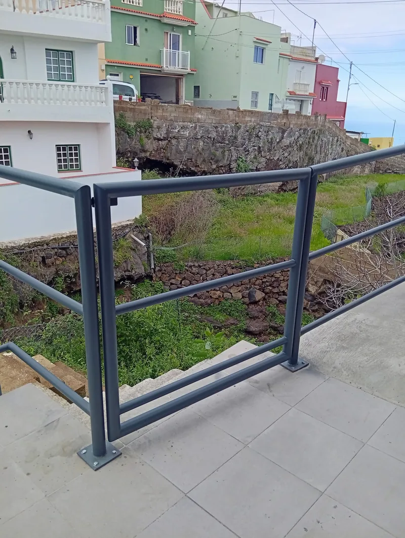 Puerta de tubos grises integrada en una barandilla de terraza junto a unos peldaños