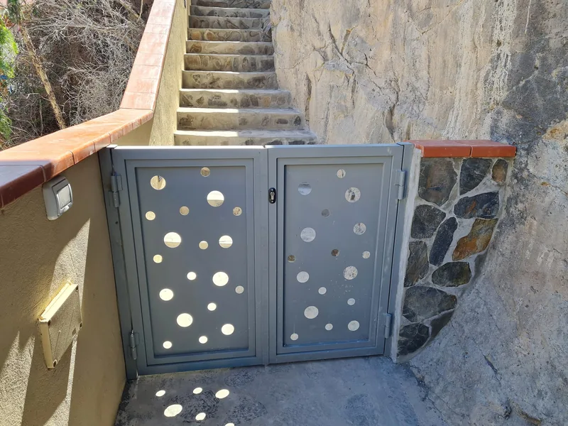 Cancela gris de dos hojas con perforaciones circulares delante de una escalera de piedra