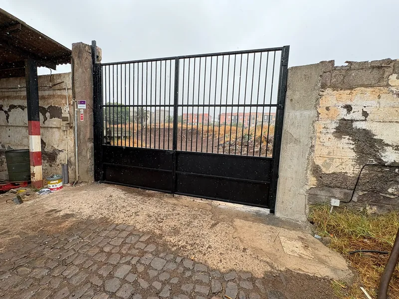 Cancela negra de dos hojas con barrotes verticales y zócalo de chapa entre muros de un acceso