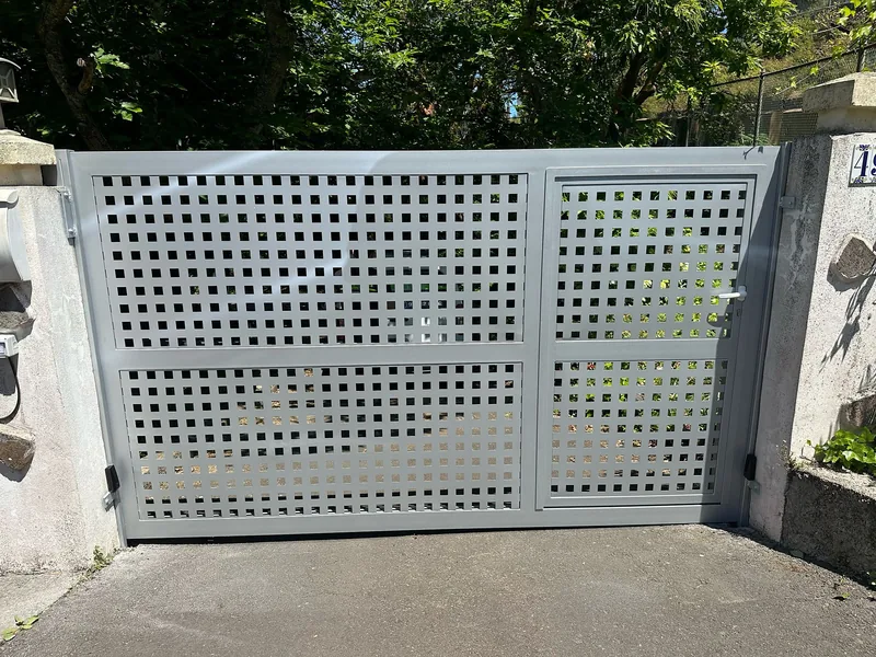 Cancela gris de perforaciones cuadradas con puerta peatonal integrada entre pilares de piedra