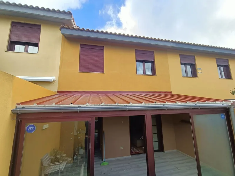 Cerramiento de perfiles oscuros y paños transparentes con cubierta rojiza junto a una vivienda