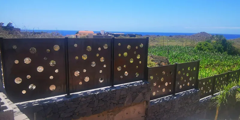 Cerramiento de paneles metálicos oscuros con agujeros circulares sobre un muro escalonado de piedra