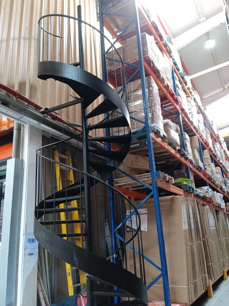 Escalera metálica de caracol negra con barandilla curva junto a estanterías de un almacén
