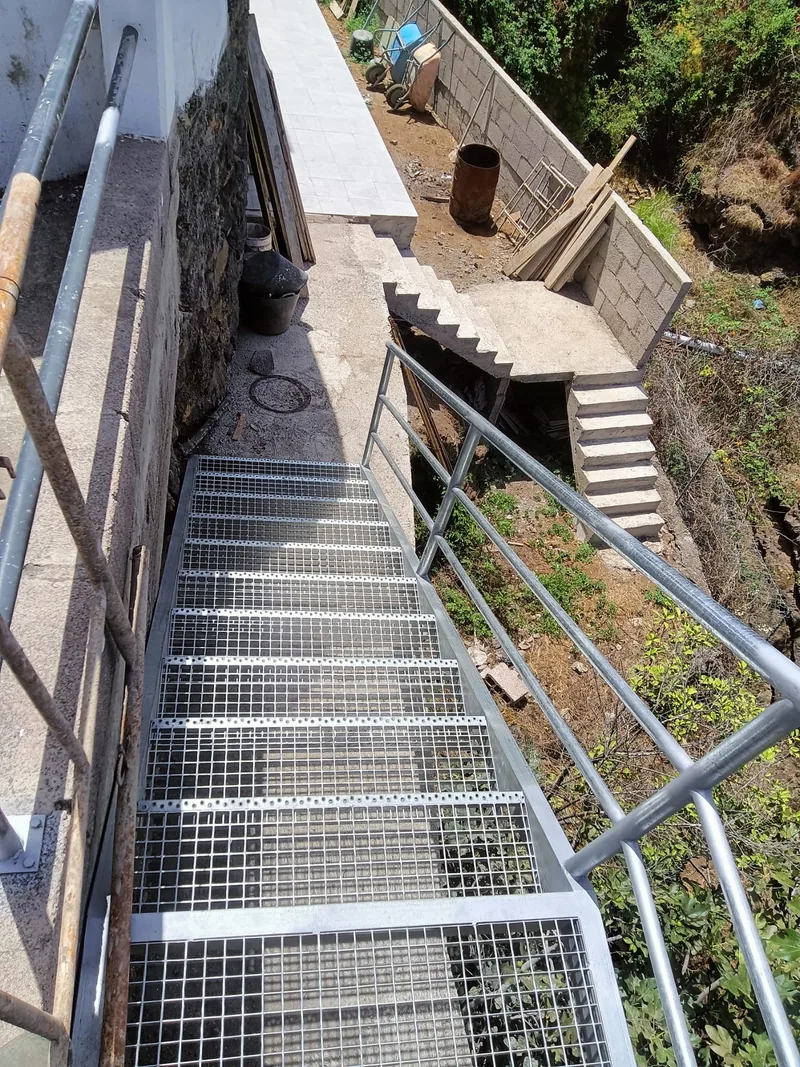 Escalera metálica exterior de peldaños de rejilla vista desde arriba junto a un muro
