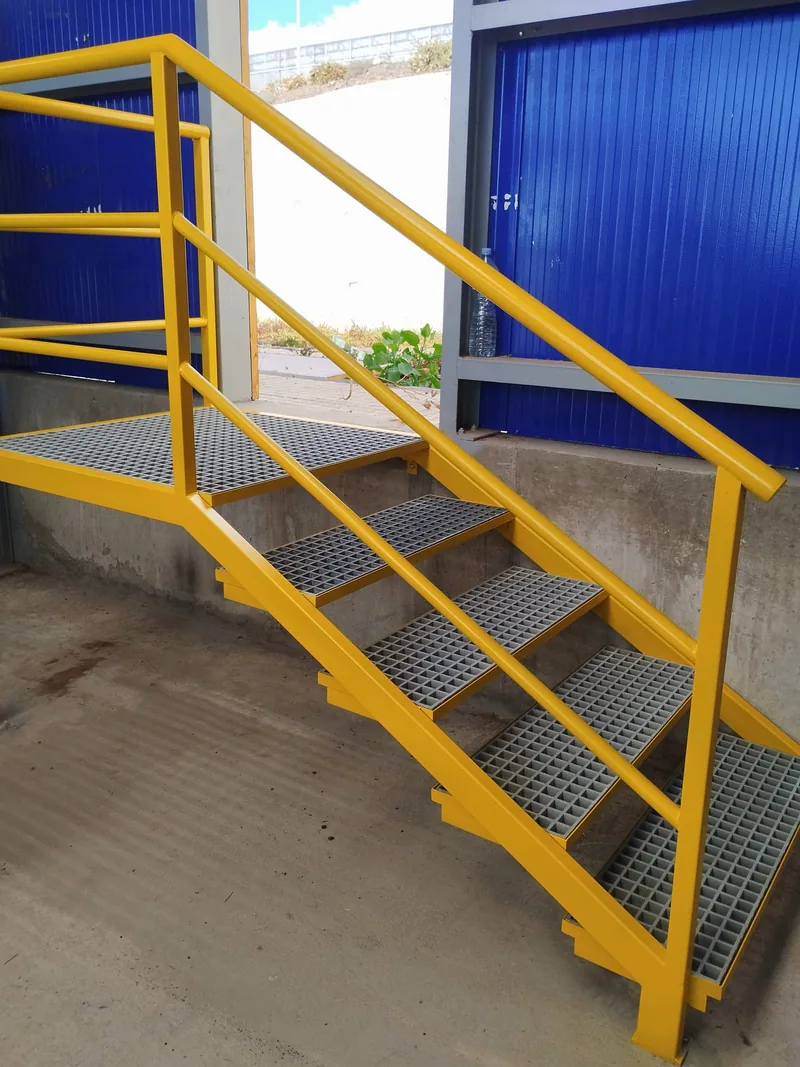 Escalera amarilla de tramo corto con peldaños de rejilla y barandilla junto a un cerramiento azul