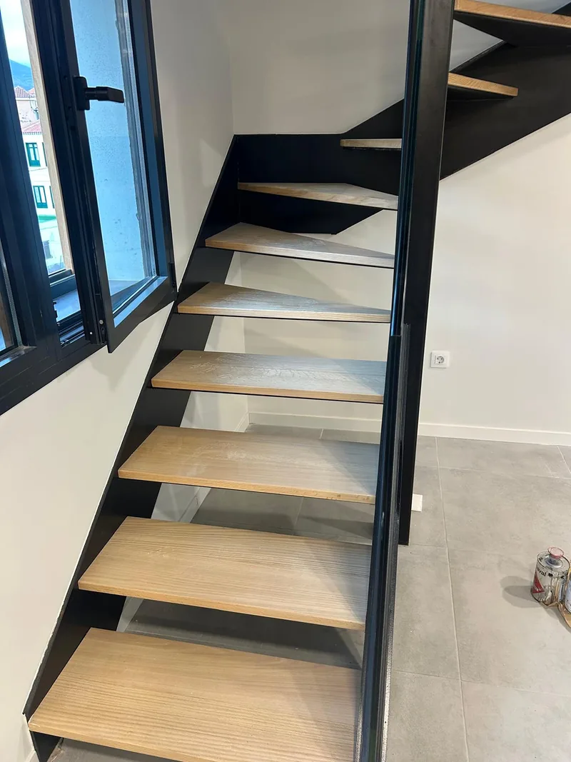 Tramo inferior de una escalera oscura con peldaños de acabado madera separados junto a una ventana