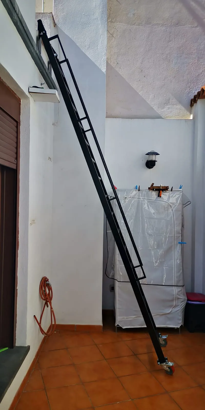 Escalera metálica negra con peldaños abiertos, agarres laterales y ruedas de apoyo junto a una pared