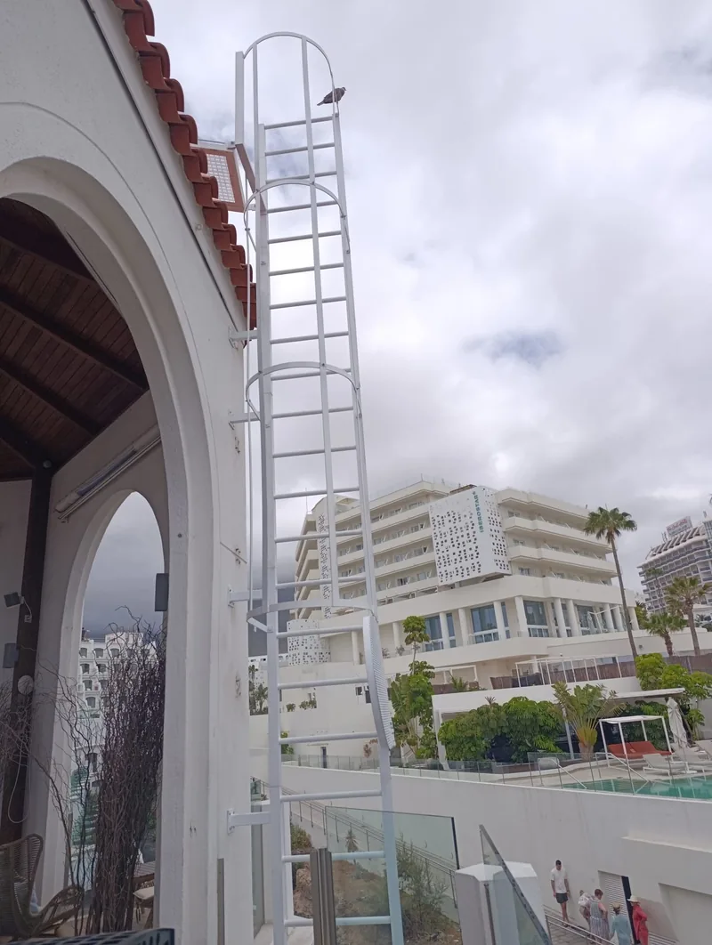 Escalera vertical blanca con aros junto a una fachada de arcos y alero de tejas