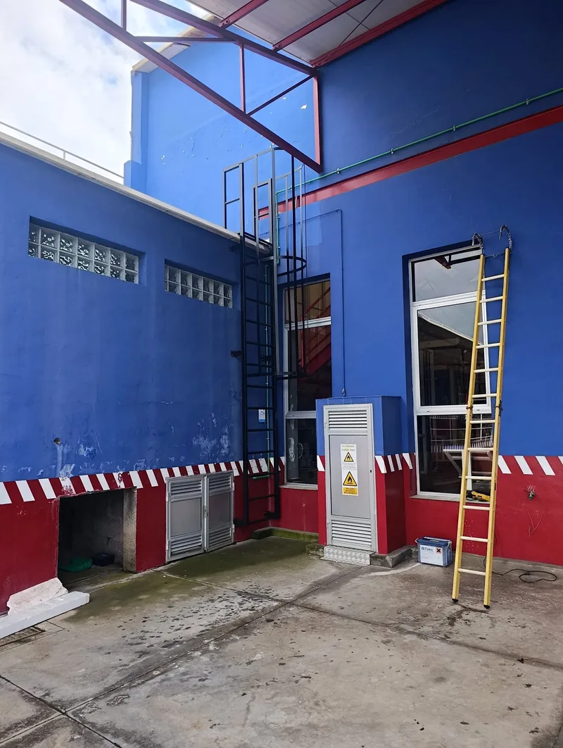 Escalera vertical oscura con aros y montantes fijada a una fachada azul en un patio industrial