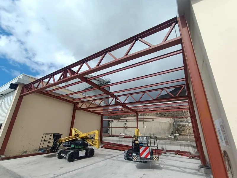 Estructura metálica rojiza de pilares, cerchas y perfiles longitudinales entre dos edificios