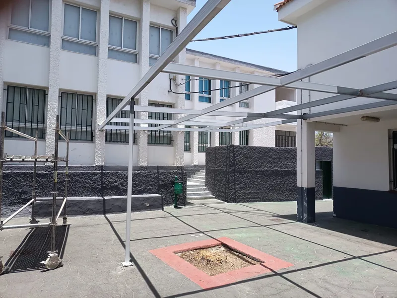 Estructura metálica de pilares y vigas sobre un patio entre fachadas blancas con zócalos oscuros