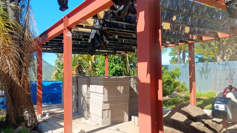 Pilares y vigas rojizas junto a una palmera con trabajos de cubierta en la parte superior