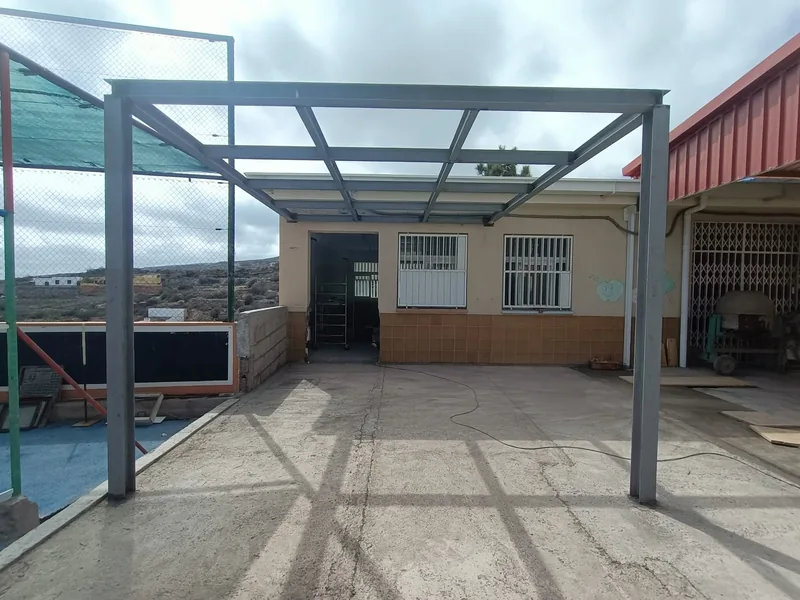 Estructura de pérgola con pilares y travesaños metálicos delante de un edificio junto a un patio