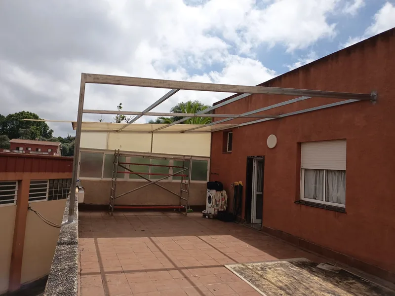 Estructura metálica de una pérgola adosada a una fachada rojiza sobre una terraza de baldosas