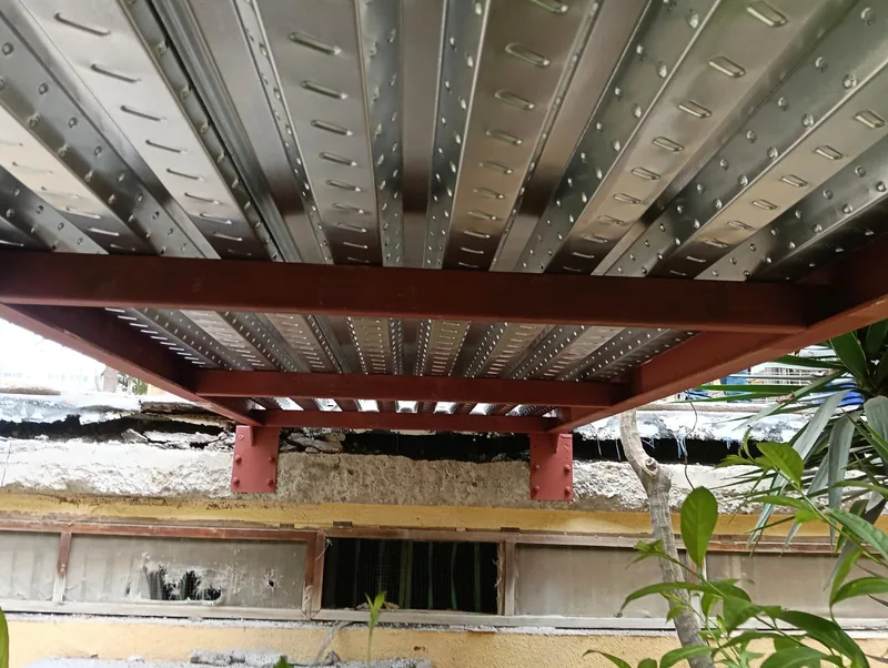 Parte inferior de chapa nervada sobre vigas rojizas y placas de unión junto a una fachada