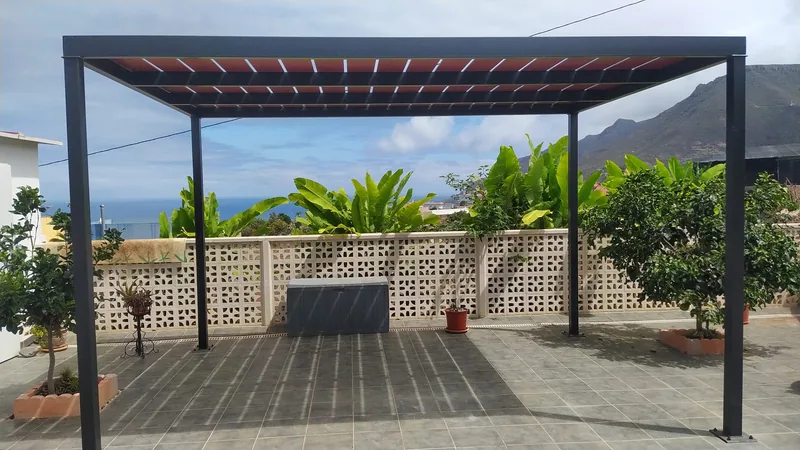 Pérgola metálica oscura con cubierta rojiza sobre una terraza de baldosas