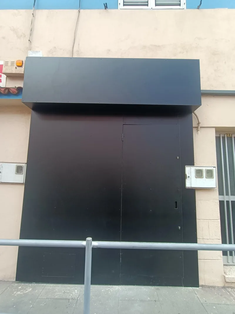 Frente metálico oscuro de panel liso con puerta peatonal integrada y remate superior ancho