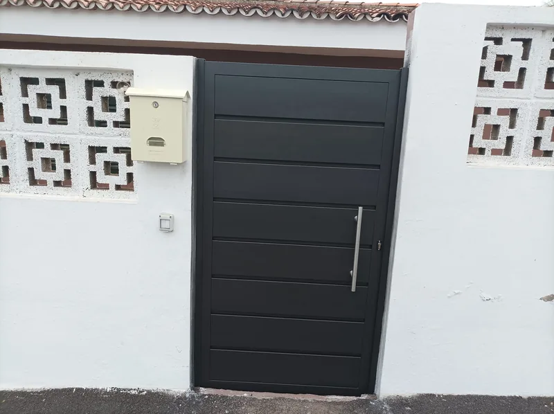 Puerta peatonal gris oscura con tirador vertical junto a un muro blanco