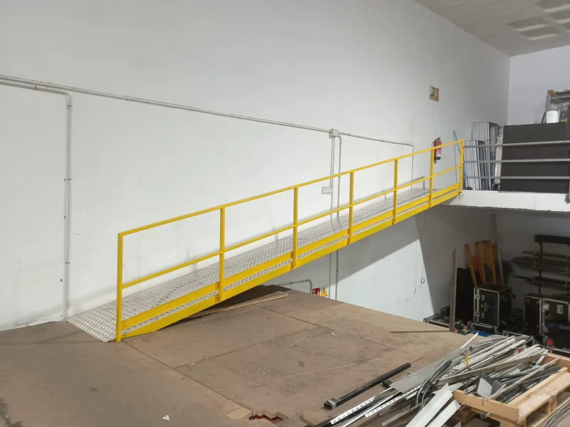 Rampa metálica inclinada con barandilla amarilla entre dos plataformas de un almacén