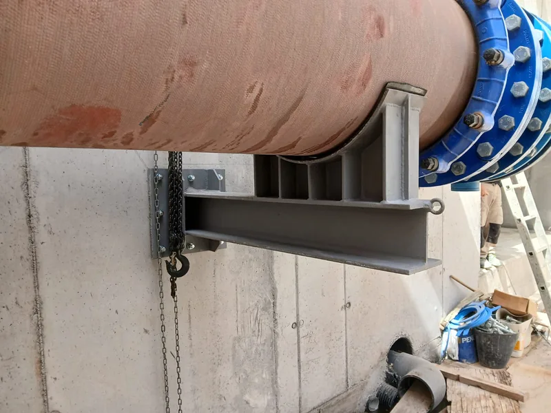 Soporte metálico fijado a un muro con placa atornillada, perfil horizontal y apoyo curvo bajo una tubería